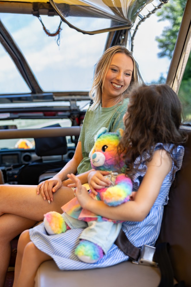 Two people sitting in a vehicle, one holding a colorful teddy bear.