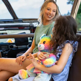 Two people sitting in a vehicle, one holding a colorful teddy bear.