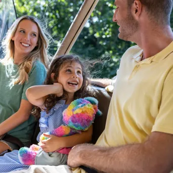 Family of three smiling in an open vehicle with greenery in background.