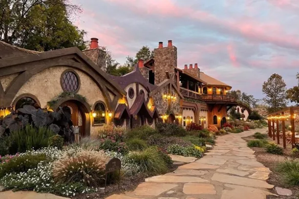 Whimsical house with arched roofs and warm lights, surrounded by gardens under a pink sky.
