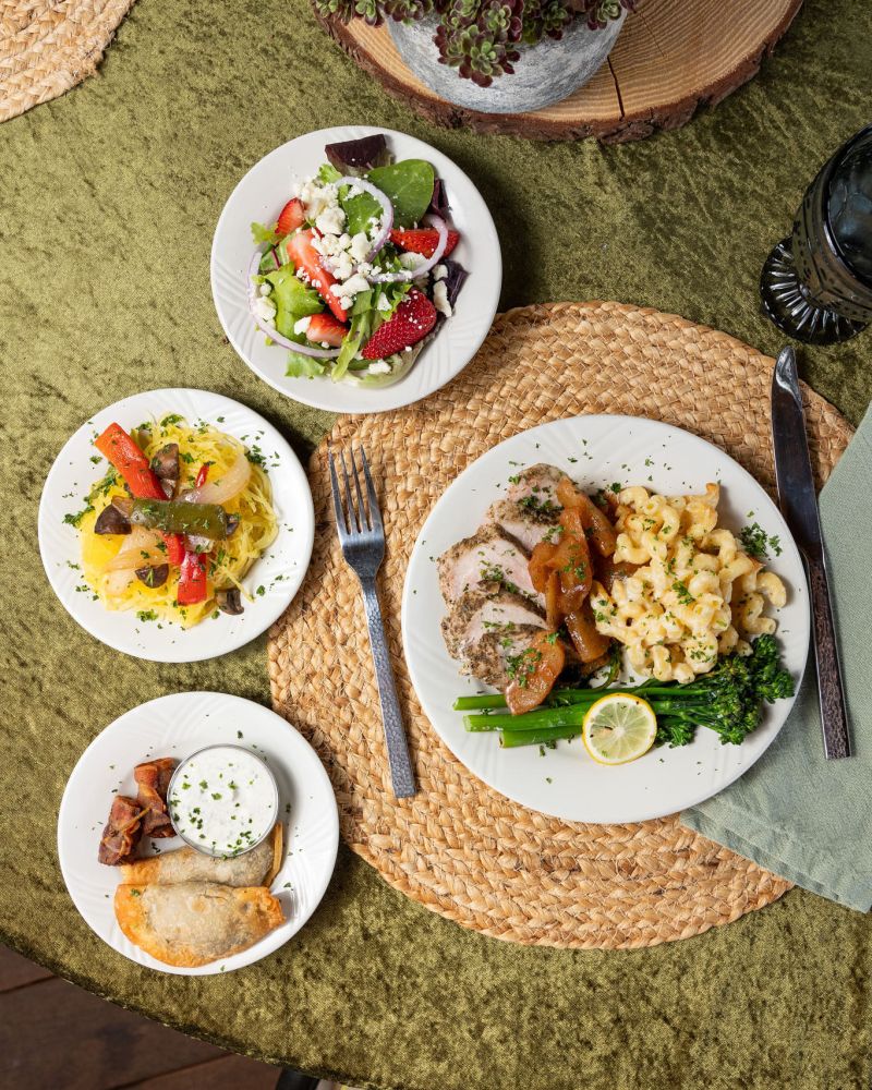 Four plates of diverse foods on a green tablecloth, featuring salad, pasta, meat, and a pastry. Ancient Lore Village Scenic Tour and Lunch with Smoky Mountains Jeep Tours.