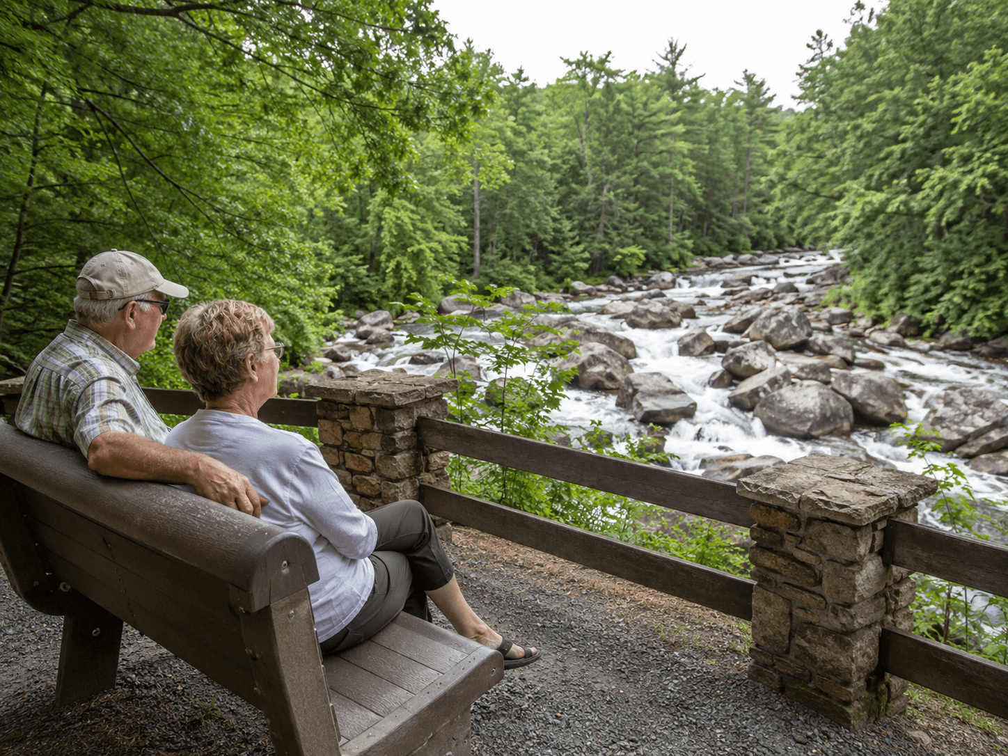 Elderly couple sitting on a bench, overlooking a rocky river with dense trees.